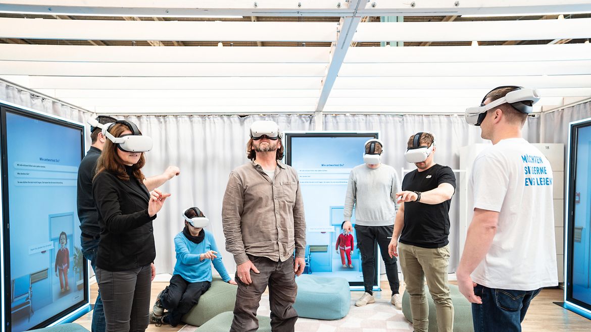 In der Ausstellung "In Würde Abschied nehmen" tragen sechs stehende Personen Virtual-Reality-Brillen, eine Person sitzt, drei menschengroße Bildschirme an den Wänden.