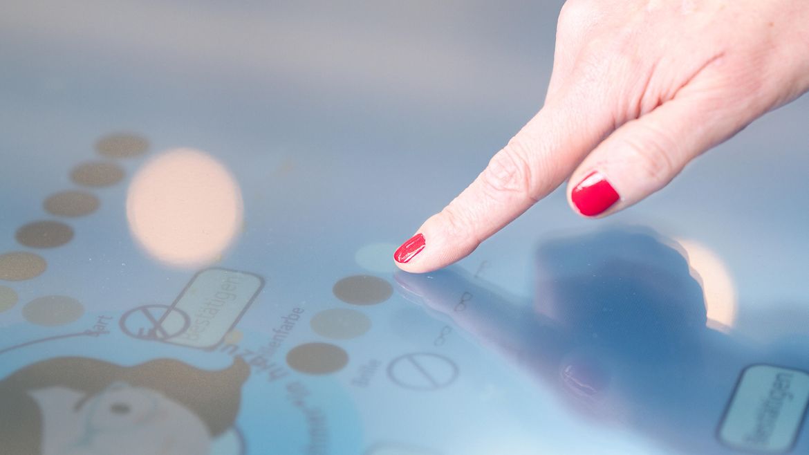 Hand mit rot lackierten Fingernägeln befindet sich über einem Bildschirm mit verschwommenem Inhalt. Die Person wählt durch Tippen einen Menüpunkt aus.