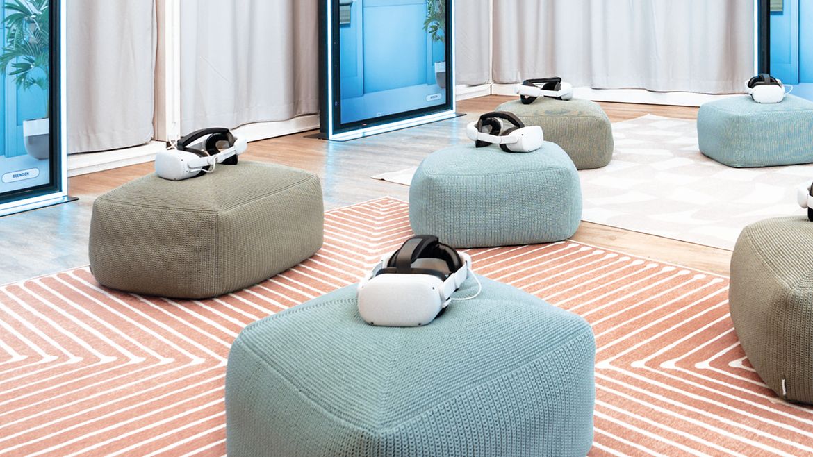 Blick in die Ausstellung "In Würde Abschied nehmen": Grüne und blaue Sitzhocker mit Virtual-Reality-Brillen darauf stehen auf pastell-orange gestreiftem Teppich.